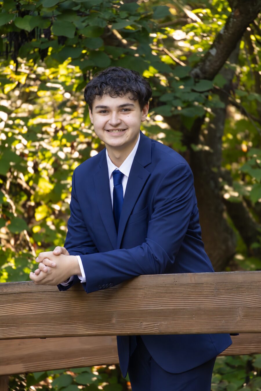 Cooper Bailey in a navy suit, smiling and leaning on a wooden railing among green trees
